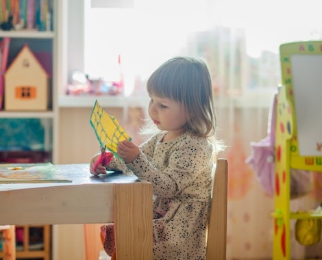 A young girl uses scissors to cut paper whilst sat at a desk.